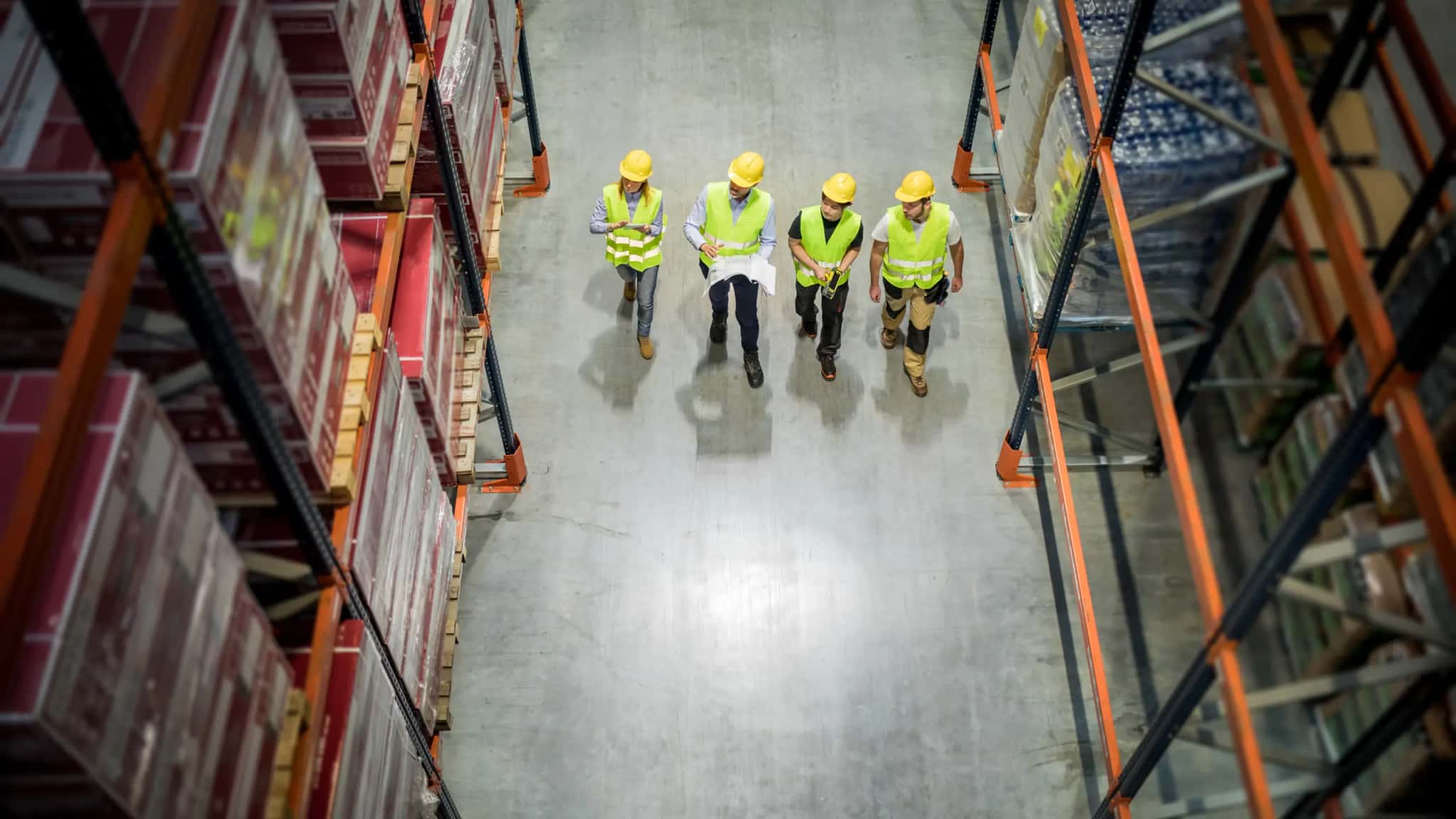 Top-down view: four workers in vests and hard hats walk down a distribution center aisle
