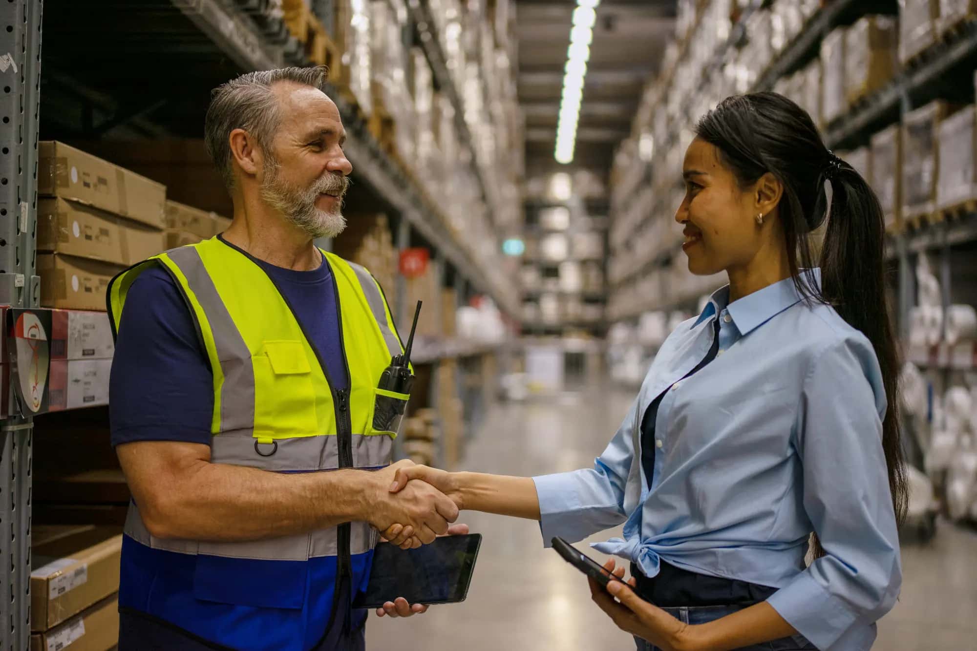 A warehouse supervisor and a manager holding a tablet shake hands between distribution center racks
